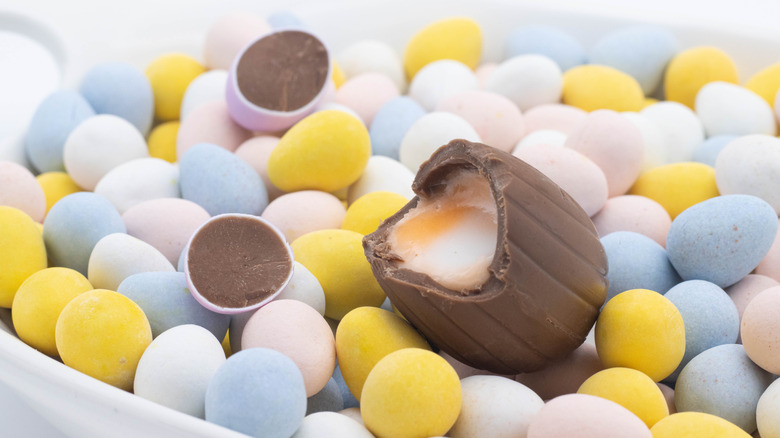 An unwrapped Cadbury Creme Egg resting on a bed of smaller candy eggs. The Cadbury egg is broken open to show the yolk-like candy filling