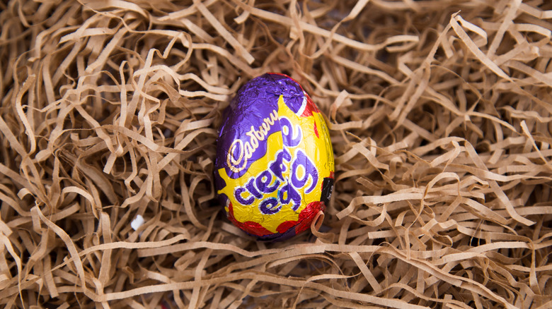 One Cadbury Creme Egg, wrapped in its red, yellow, and purple foil, on a nest of brown paper strips