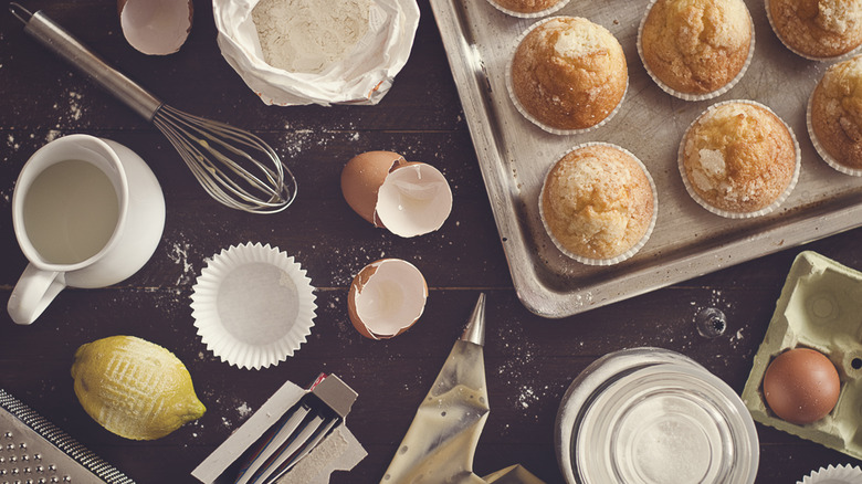 A countertop covered in tool and ingredients for making muffins such as a whisk, milk, muffin cups, a lemon, and a piping bag next to finished muffins