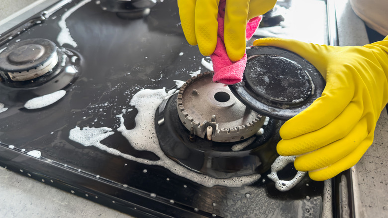 Yellow-gloved hands cleaning black gas stove burners with a pink rag and soapy solution