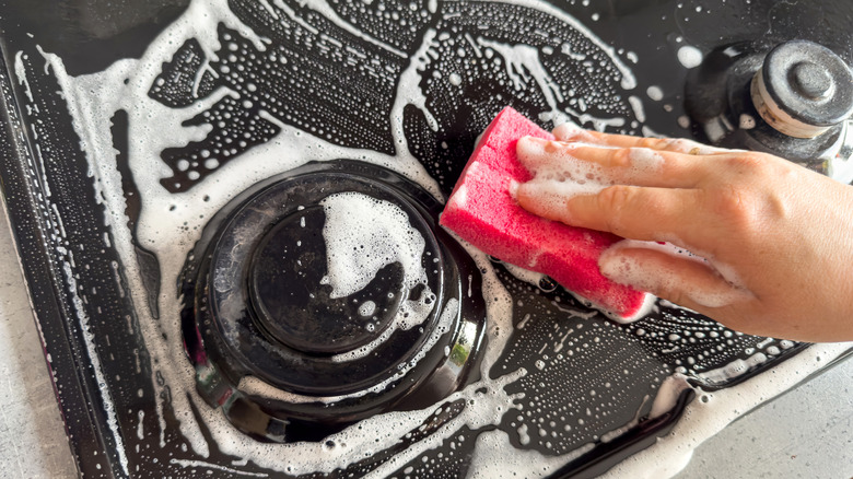 Cleaning a gas stove-top surface with a soapy cleaner and pink sponge