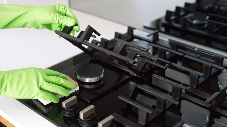 Green-gloved hands lifting a grate on a gas stove-top to wipe underneath it with a sponge