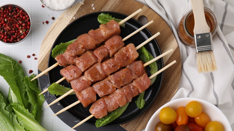Skewers of raw pork marinating on dish, marinade and brush beside.