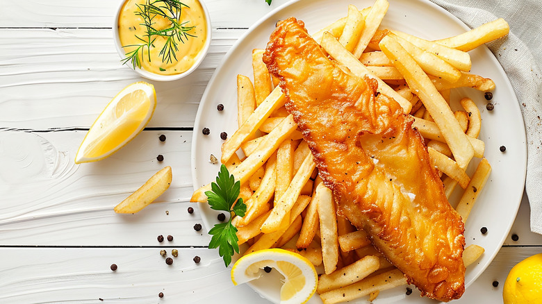 A large piece of fish on top of french fries on a white plate with lemon slices and sauce