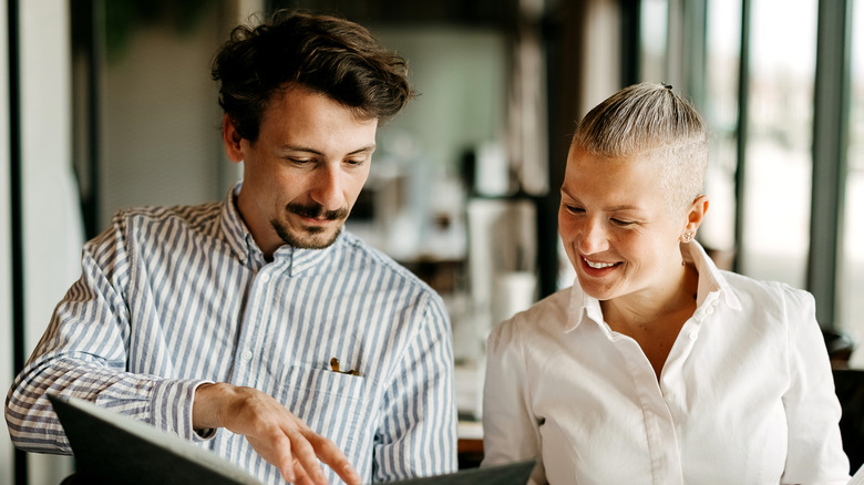 A well-dressed couple sits at a restaurant and looks at the menu with glasses on the table in front of them