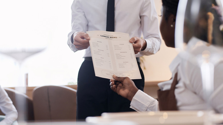 A server in a shirt and tie hands someone else a lunch menu