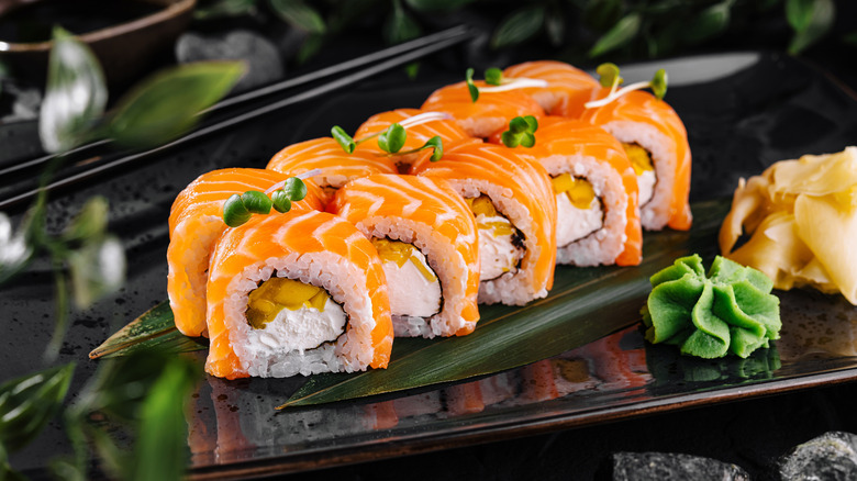 Salmon sushi roll on dark plate by garnishes and chopsticks