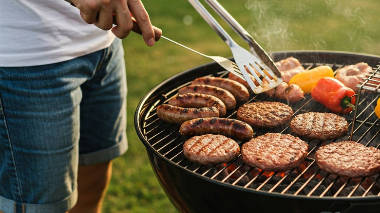Close up of a person using a grill that's covered with hamburgers, sausages, and peppers