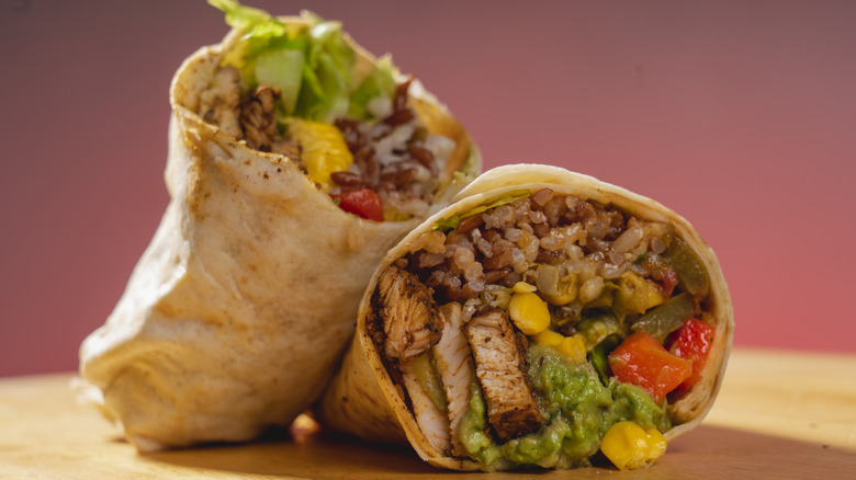A burrito sliced in half, showing the filling of chicken, rice and beans, peppers, lettuce, guacamole, and corn.