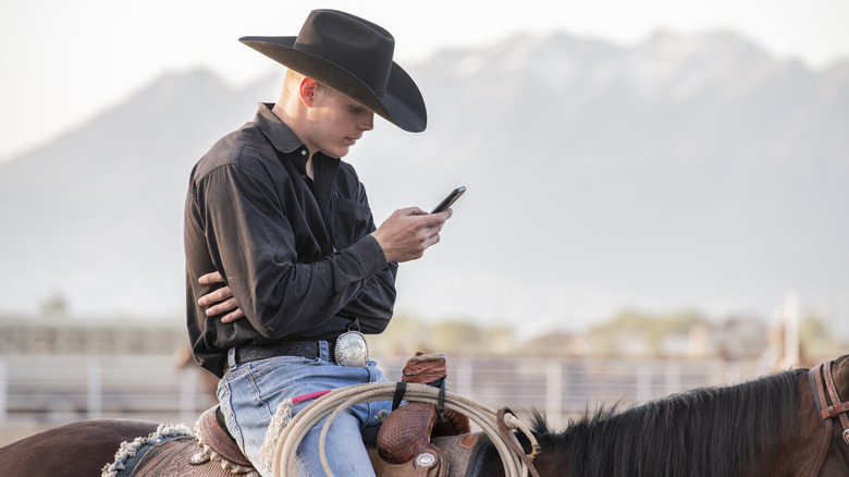 A man in a cowboy hat, sitting on a horse and checking his phone