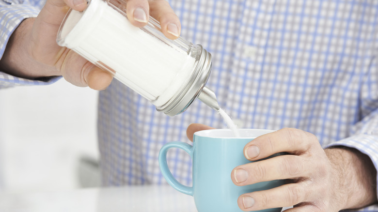 Person pouring sugar from a dispenser into a mug of coffee
