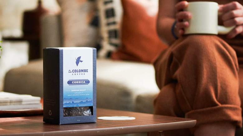 A box of La Colombe Corsica sitting on a coffee table with person holding a coffee cup in the background