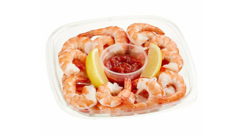 a plastic container of shrimp with a ramekin of cocktail sauce and sliced lemons