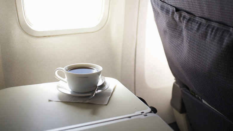 A white cup of black coffee on an airplane seat tray, in the window light
