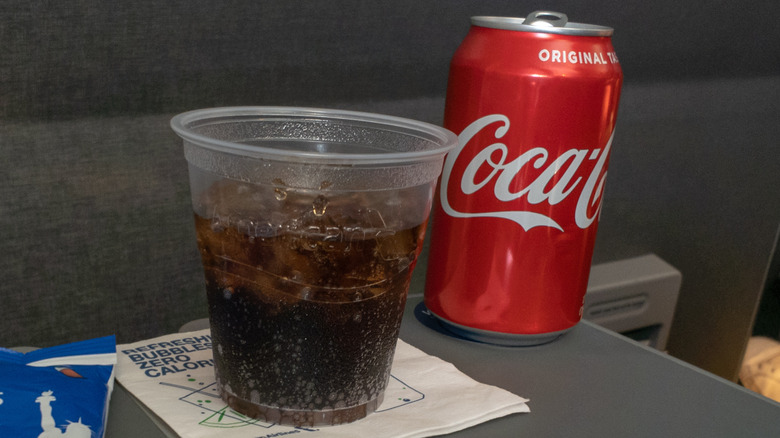 A can of Coca-Cola next to a plastic cup filled with the same, on an airplane seat tray