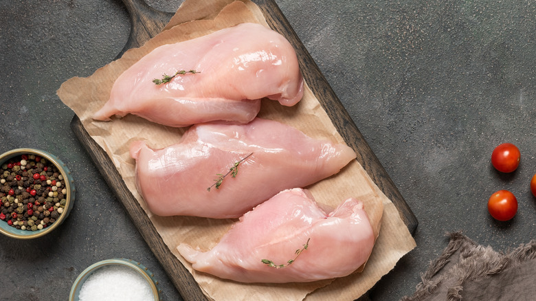 Raw chicken breasts on a wooden board with cherry tomatoes, pepper, and salt on the side