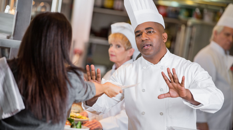 A chef yelling at a restaurant server who's handing him a ticket