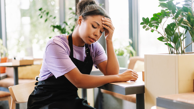 A young restaurant waitress sits at an empty table looking stressed and tired