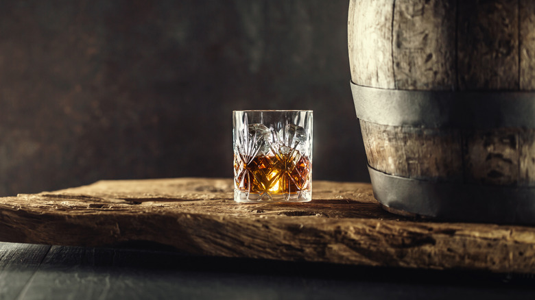 Glass of bourbon in ornamental glass next to a vinatge wooden barrel on a rustic wood and dark background.
