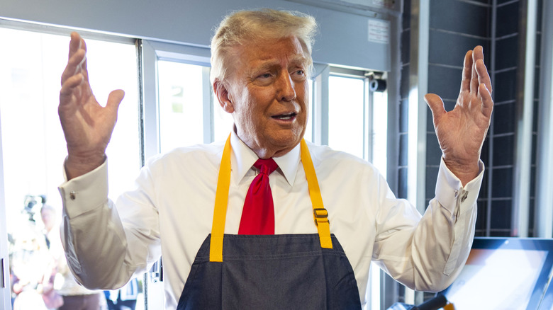 President Donald Trump inside a fast food restaurant drive thru wearing an apron with is hands in the air.
