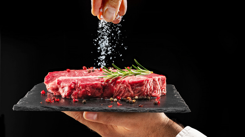Close-up of a stone slab with a steak and seasoning being sprinkled onto it