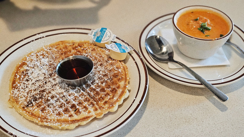 A plate with a Waffle House waffle topped with powdered sugar, a ramekin of syrup, and two packets of butter next to a cup of tomato soup