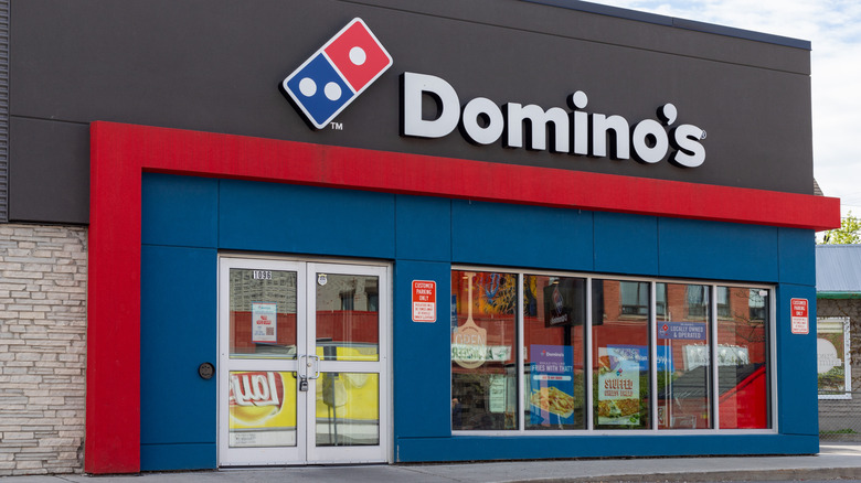 The entrance to a Domino's pizza restaurant with its classic red and blue domino sign and advertisements for stuffed pizza and fries in the window