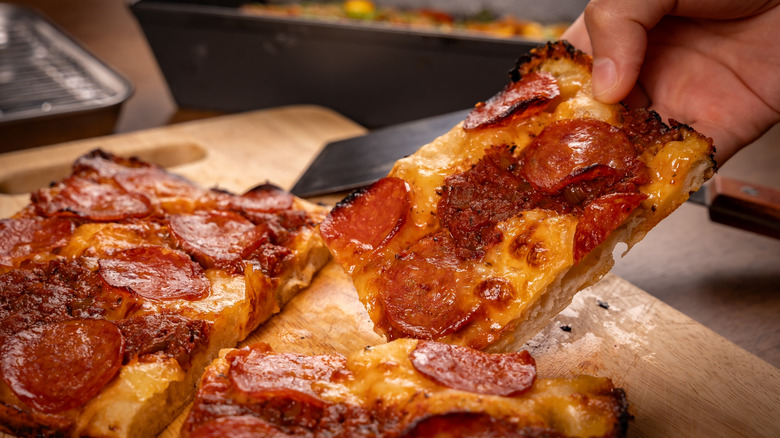 A hand taking a slide of Detroit-style pizza from a wooden board