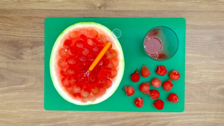 Half watermelon with balls removed and glass of juice on green cutting board