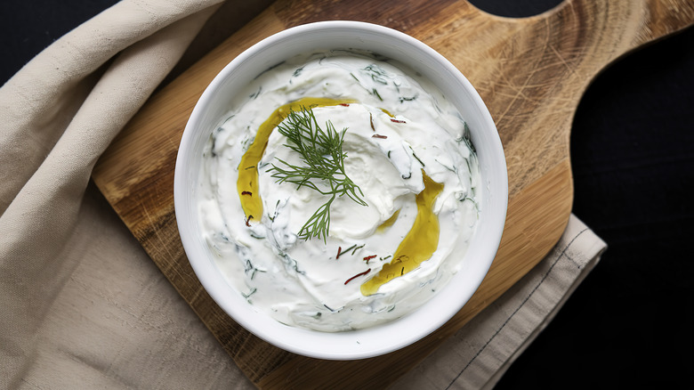 Tzatziki in a white bowl with sprig of fresh dill, sliced cucumbers.