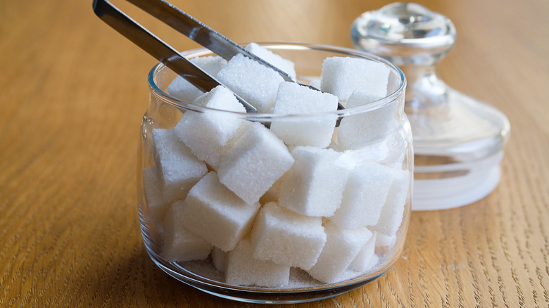 A glass bowl of sugar cubes with metal tongs on a wooden table