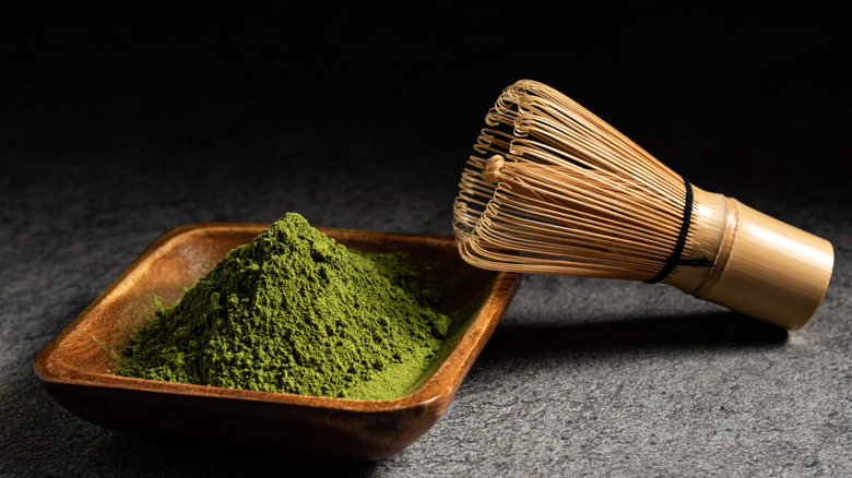 Matcha bamboo whisk resting on a square wooden bowl with a pile of powdered matcha