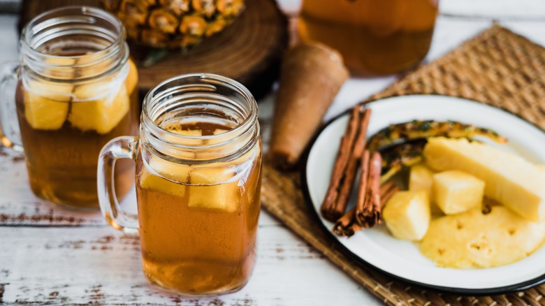 Two mugs of tepache by plate of pineapple and cinnamon