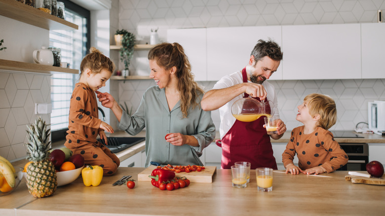Family of four in kitchen with two children at the counter having orange juice and cutting vegetables