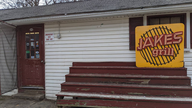 The humble exterior of Jake's Grill sports a yellow sign, worn wooden bleacher seats and white siding on a small country house building.