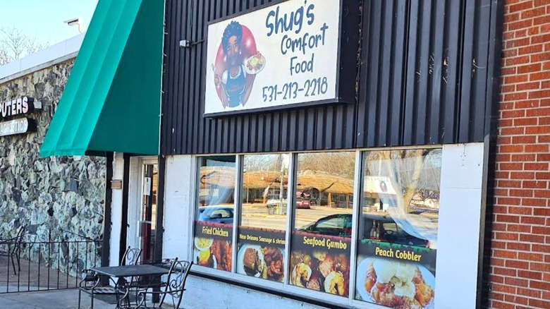 The outside of Shug's Comfort Food in Bellevue, Nebraska sports a sign with an artistic imagination of the chef, as well as window decor advertising their fried chicken, seafood gumbo and peach cobbler.