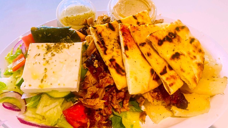 A chicken souvlaki platter from Jersey Gyro is piled with a feta block, warm pita bread, red onion, tomatoes, and lettuce.