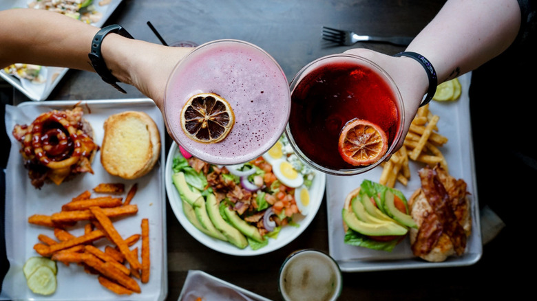 People toasting with blood orange cocktails above plates with a selection of foods on a table in Koury's Tavern.