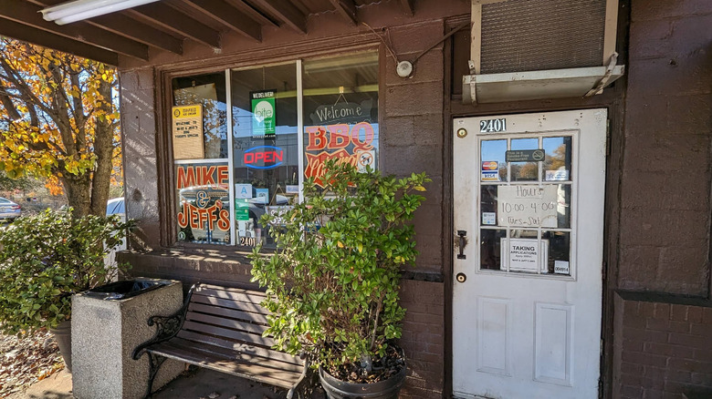 A bench sits in front of the simple entrance to Mike and Jeff's BBQ in Greenville, South Carolina, next to a window with handpainted signage.