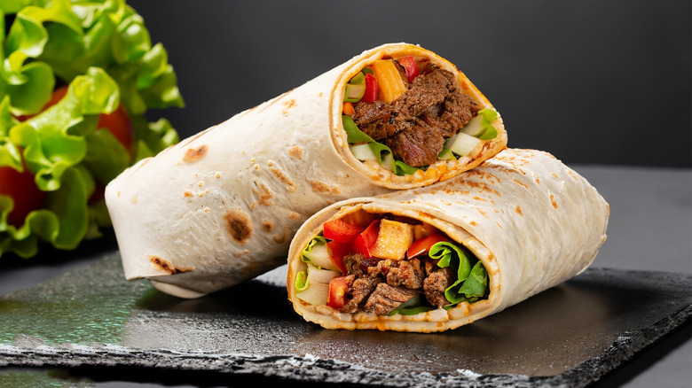 a delicious looking grilled carne asada burrito cut in half sitting on a black slate board