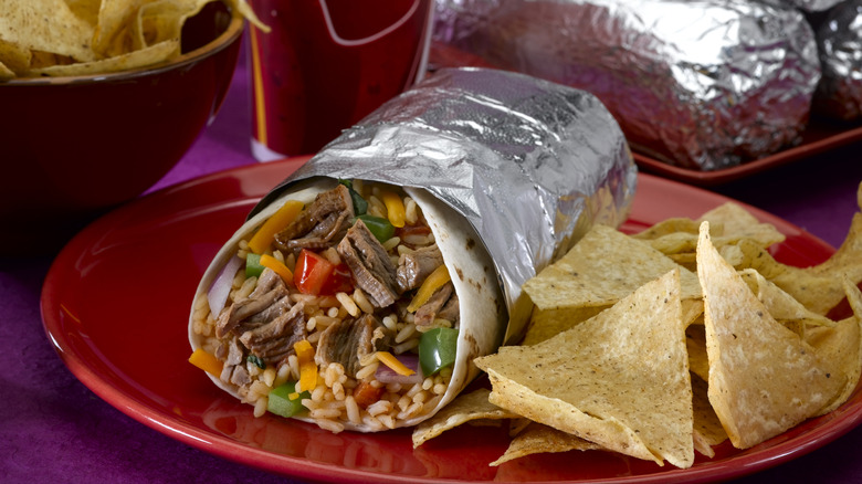 a burrito wrapped in foil on a red plate with a stack of foil-wrapped burritos in the background
