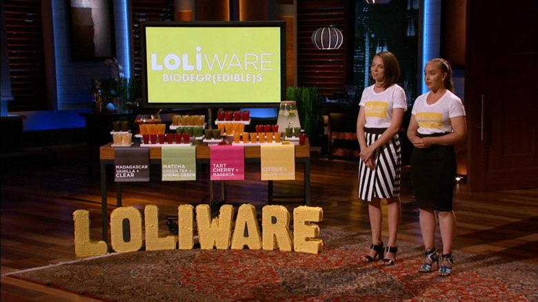 Two women standing in white t-shirts and black/black and white dresses next to a table of colored cups, labeled with flavors and colors, and the logo 