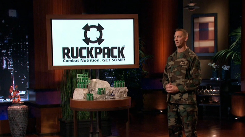A man in green army camo stands next to a table of green energy shot bottles, with the logo for 