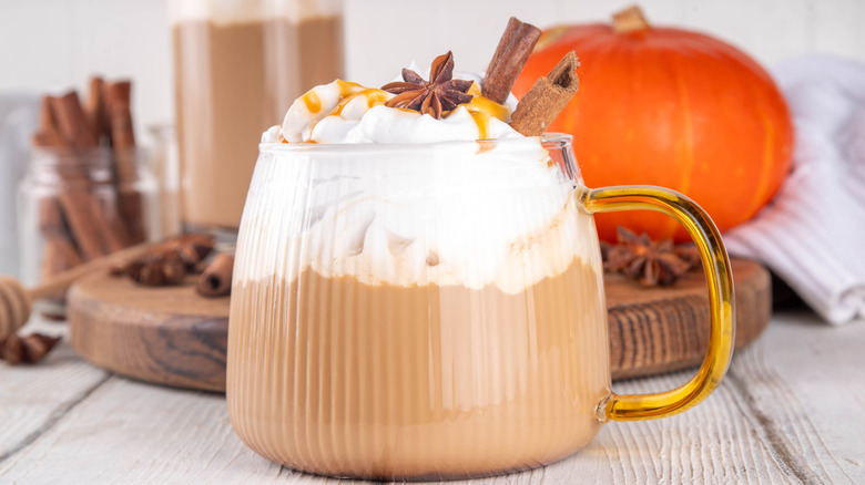Glass mug of pumpkin spice latte topped with whipped cream, pumpkin pie syrup, anise, and cinnamon sticks with an orange pumpkin and spices in the background