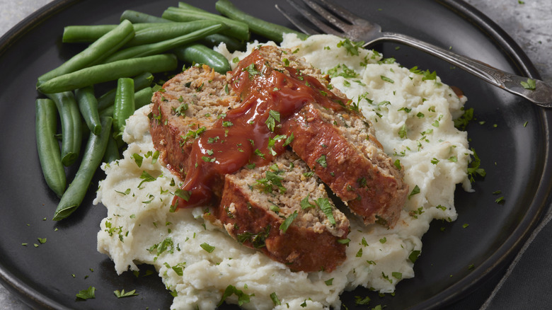 Meatloaf over mashed potatoes with a side of green beans on a black plate