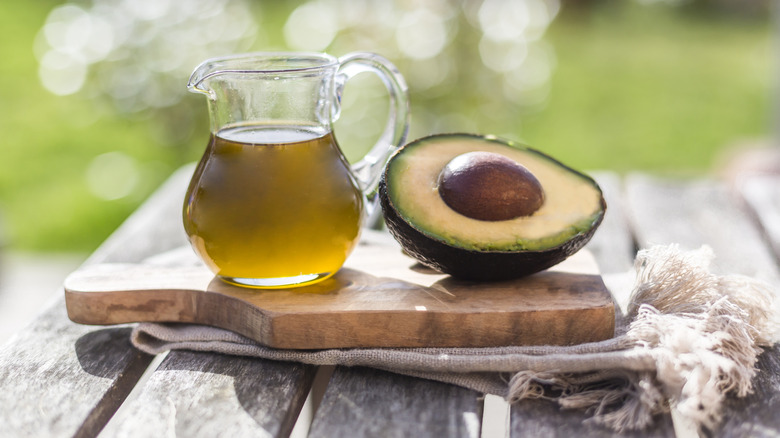 Glass pitcher of avocado oil and half of an avocado on a wooden board