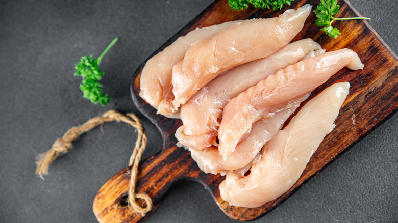 Raw chicken tenderloins laid on a wood cutting board with some herbs