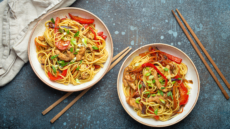 Two plates of chicken lo mein with green onion and red peppers, and chopsticks off to the side