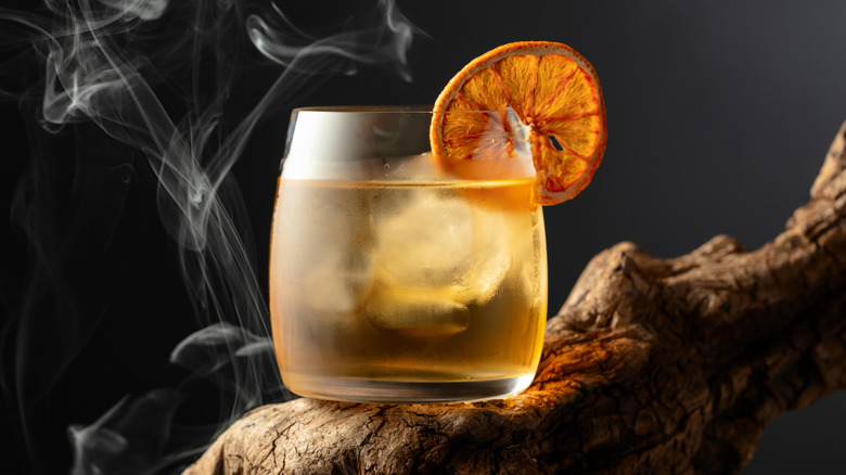 Rum cocktail with ice and orange garnish with smoke in the background on a black backdrop