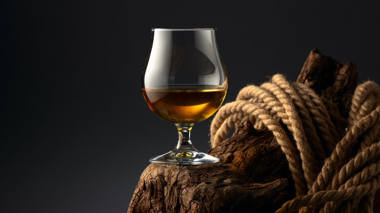 Glass of rum cut with water to make grog on a wooden log with rope against a dark background